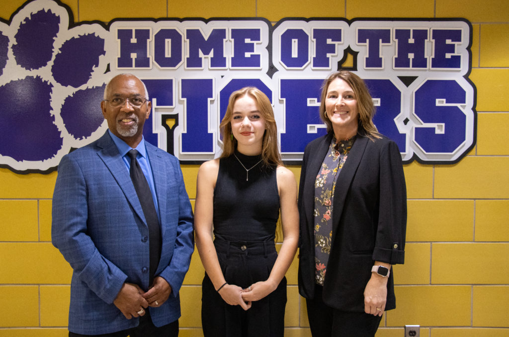 A group of three people standing in front of a yellow brick wall with a sign that reads 'Home of the Tigers.' In the center is a young woman wearing a black sleeveless top and black pants. To her left is a man in a blue plaid suit and black tie, and to her right is a woman wearing a black blazer over a floral blouse. All three are smiling at the camera.