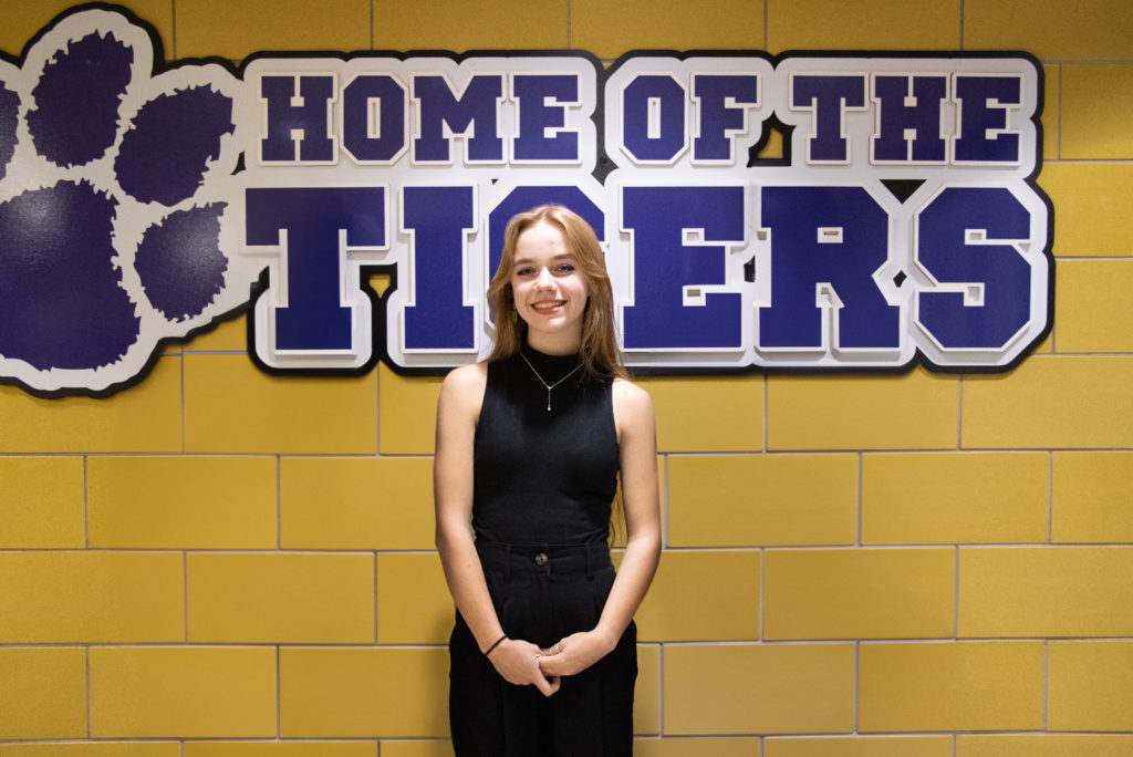 A young woman standing in front of a yellow brick wall with a large sign that reads 'Home of the Tigers.' She is wearing a black sleeveless top and black pants, smiling at the camera.