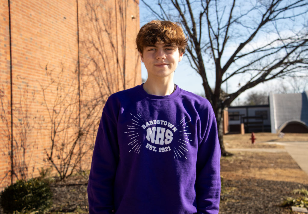 A young person with light skin and light brown hair stands outside in daylight, smiling softly at the camera. They are wearing a purple Bardstown NHS (National Honor Society) sweatshirt, which has white text and a burst design around the logo. The background features a brick building, leafless trees, and a clear blue sky, suggesting a late fall or winter season. The setting is outside the Bardstown Polytechnic Center.